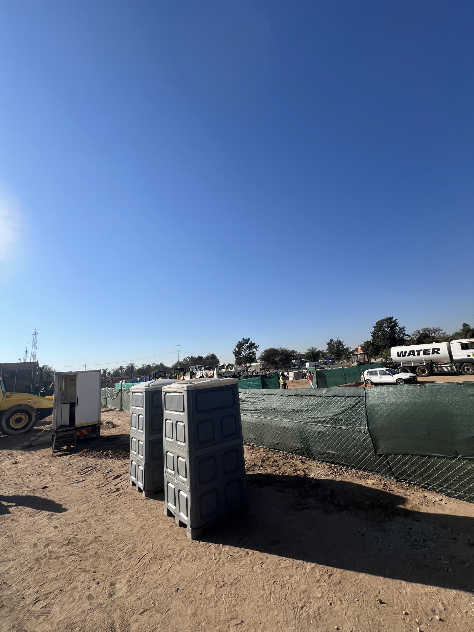 Two chemical toilets on pallets beside shade-cloth site fencing, with a water tanker and plant working on a construction site near Polokwane